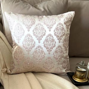 May include: A square throw pillow featuring a cream-colored satin fabric with a repeating dusty pink damask pattern. The design is accented with small, clear, reflective beads embedded within the intricate floral motifs across the surface.