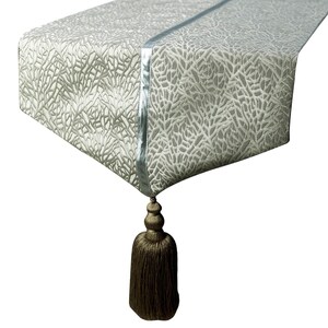 May include: A rectangular table runner featuring a light grey and white textured coral-like pattern across the surface. The runner includes a smooth, solid light grey central strip and a large, dark brown decorative tassel hanging from the end.