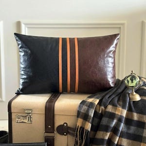 May include: A rectangular throw pillow featuring a color-blocked design with a black left side and a dark brown right side, separated by three vertical tan stripes. The pillow has a smooth, leather-like texture and a clean, structured appearance.