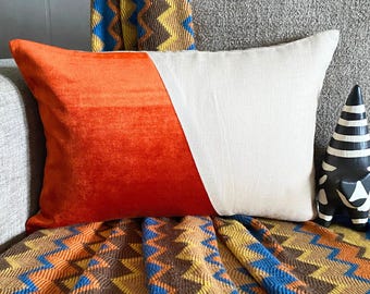 May include: A rectangular throw pillow featuring a color-blocked design with one side in a vibrant orange velvet fabric and the other side in a plain off-white linen-like material, divided by a diagonal seam across the center of the cushion.
