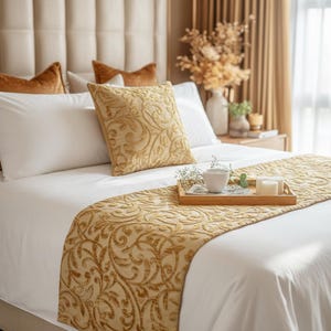 Puede incluir: Una cama con funda nórdica blanca, un camino de cama dorado con un intrincado patrón de volutas en relieve y un cojín cuadrado a juego. Una bandeja de madera sobre la cama sostiene una taza de cerámica blanca y dos velas blancas.