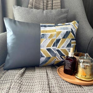 May include: A rectangular throw pillow featuring a solid dark grey panel on the left and a patterned panel on the right with a geometric chevron design in mustard yellow, dark blue, and white. A brown glass bottle of Estée Lauder serum sits nearby.