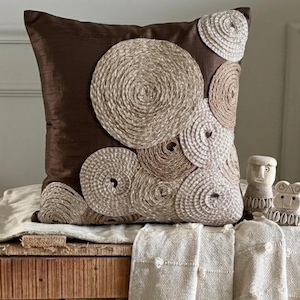May include: A square throw pillow with a dark brown fabric base featuring a cluster of beige and tan circular rope appliques of varying sizes arranged across the surface. The textured rope coils create a raised, spiraling pattern on the pillow.