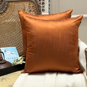 May include: Two square, rust-colored throw pillows with a textured appearance. The pillows are placed on a chair with a white blanket. A book and a tea set are visible in the background, suggesting a cozy interior decor theme.