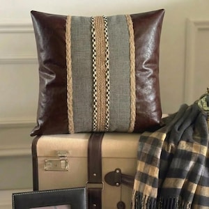 May include: A square throw pillow featuring dark brown faux leather side panels and a central gray fabric panel. The design includes two vertical braided jute ropes flanking a central woven strip with a repeating geometric pattern in tan and black.