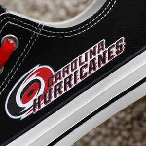 May include: Black canvas shoe with white trim and red laces. The Carolina Hurricanes logo is prominently displayed on the side in red, white, and black. The text "CAROLINA HURRICANES" is also visible.