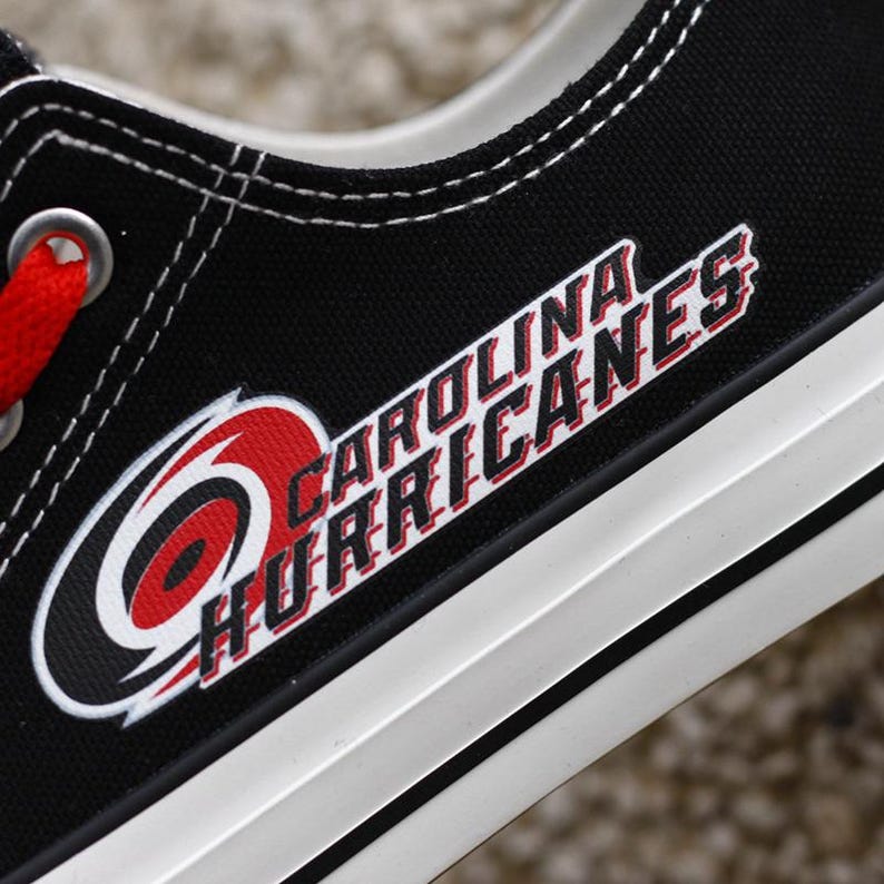 May include: Black canvas shoe with white trim and red laces. The Carolina Hurricanes logo is prominently displayed on the side in red, white, and black. The text "CAROLINA HURRICANES" is also visible.