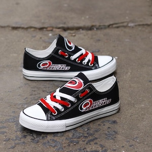 May include: Black canvas sneakers with white soles and red laces. The sneakers feature the Carolina Hurricanes logo in white and red. The shoes have a low-top design and are suitable for casual wear.