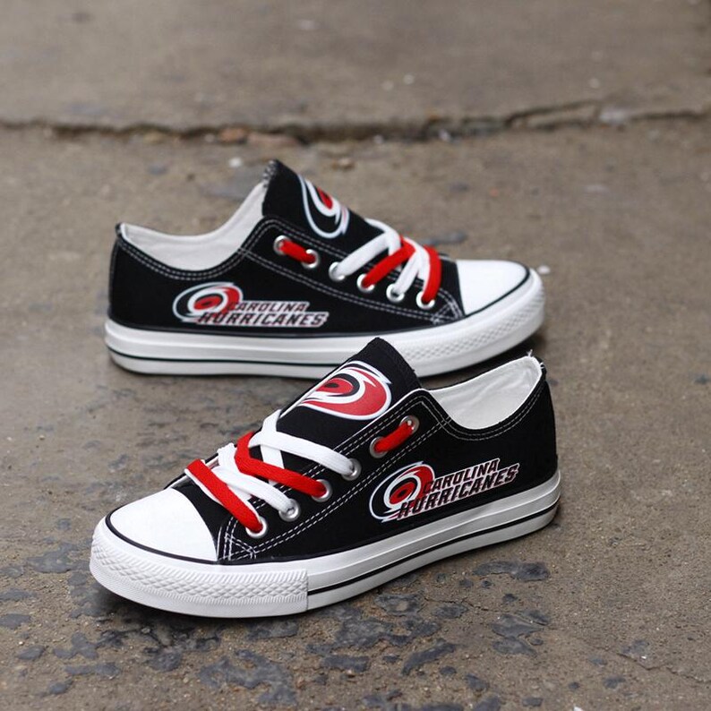 May include: Black canvas sneakers with white soles and red laces. The sneakers feature the Carolina Hurricanes logo in white and red. The shoes have a low-top design and are suitable for casual wear.
