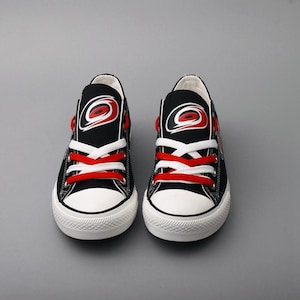 May include: Pair of black canvas sneakers with white soles and red laces. Each shoe features a logo with a red and white design on the tongue. The sneakers are designed for casual wear and have a classic, sporty look.