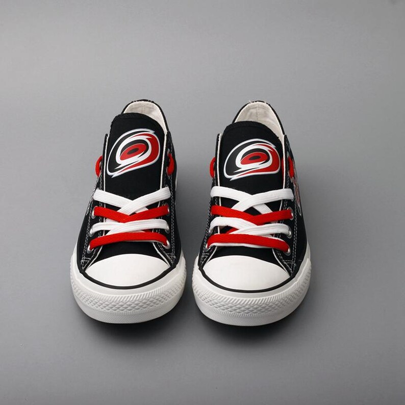 May include: Pair of black canvas sneakers with white soles and red laces. Each shoe features a logo with a red and white design on the tongue. The sneakers are designed for casual wear and have a classic, sporty look.