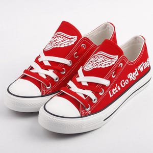 Könnte beinhalten: Ein Paar rote Canvas-Schuhe mit weißen Schnürsenkeln und einem weißen Logo eines roten Flügels mit dem Text "Let's Go Red Wings" auf einem Schuh.