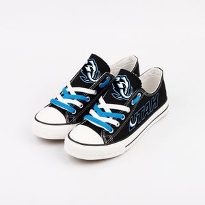 Puede incluir: Un par de zapatillas de lona negras con suelas y punteras blancas. Las zapatillas tienen cordones azules y un logotipo con la palabra "UTAH" en blanco. El logotipo es un diseño estilizado en azul y blanco.