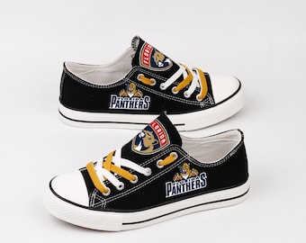 May include: Pair of black canvas sneakers with white soles and toe caps. The sneakers feature the Florida Panthers logo on the sides and tongues, with white laces and yellow laces. The shoes are designed for casual wear.