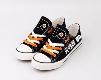Pode incluir: Um par de tênis de lona pretos com solas e biqueiras brancas. Os tênis apresentam cadarços laranja e o logotipo do Philadelphia Flyers na lateral e na língua. O logotipo é um "P" laranja e branco estilizado com asas.