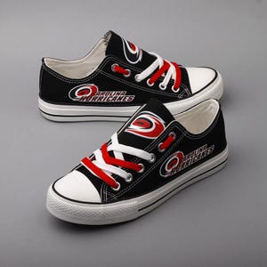 May include: Pair of black canvas sneakers with white soles and toe caps. The sneakers feature red laces and the Carolina Hurricanes logo in red and white. The shoes are displayed on a gray surface.