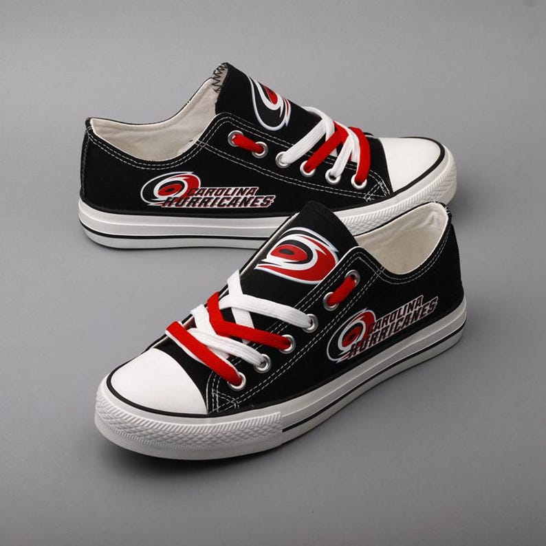 May include: Pair of black canvas sneakers with white soles and toe caps. The sneakers feature red laces and the Carolina Hurricanes logo in red and white. The shoes are displayed on a gray surface.