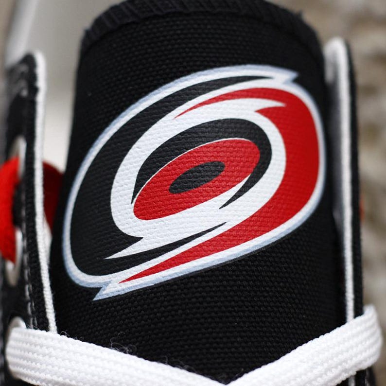 May include: Close-up of a black canvas sneaker featuring the Carolina Hurricanes logo. The logo is a red and white stylized hurricane symbol on a black background. White laces and a red lace are visible.