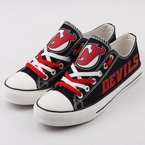 Puede incluir: Par de zapatillas de lona negras con suelas y punteras de goma blancas. Las zapatillas tienen cordones rojos y el logotipo de los New Jersey Devils en los laterales. La palabra "DEVILS" está impresa en rojo en el exterior de cada zapato.