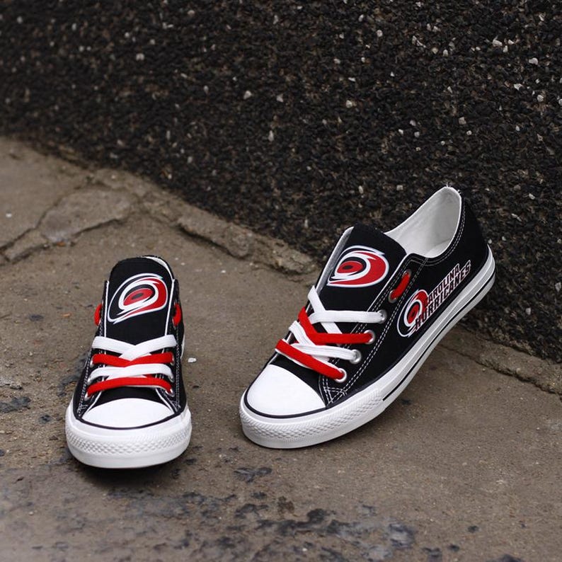May include: Black canvas sneakers with white soles and red laces. The sneakers feature the Carolina Hurricanes logo in red and white. The shoes are displayed on a concrete surface.