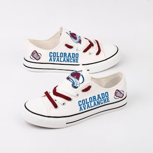 Puede incluir: Zapatillas de lona blancas con cordones burdeos y el logotipo de los Avalanche de Colorado. Los zapatos presentan el nombre del equipo en letras azules en el lateral, con una franja negra a lo largo de la suela.