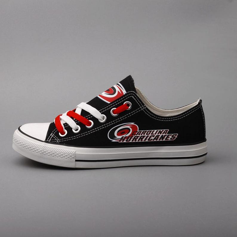 May include: Black canvas low-top sneaker with white toe cap and sole. Features red laces and the Carolina Hurricanes logo in red and white. The team name is printed on the side of the shoe.