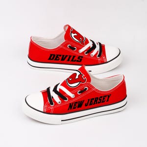 Könnte beinhalten: Rote und weiße Canvas-Sneaker mit schwarzen Schnürsenkeln und dem Logo der New Jersey Devils. Die Worte "Devils" und "New Jersey" sind auf den Schuhen aufgedruckt.