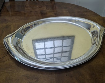 May include: A silver oval serving tray with a slightly raised rim and two curved handles. The tray is highly polished and reflects the surrounding environment.