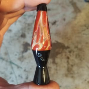 May include: A red and white lava lamp shaped glass pipe with a black base. The pipe is signed with a black letter "S".
