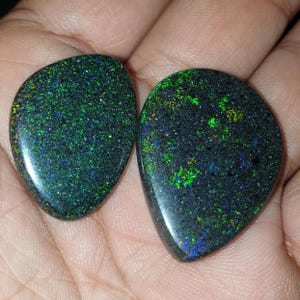 Puede incluir: Dos piedras preciosas de ópalo negro pulido con vibrantes destellos verdes, azules y amarillos. Los ópalos tienen un aspecto moteado y tienen forma de lágrima, mostrando un juego de color natural e iridiscente.