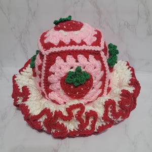 K&ouml;nnte beinhalten: Eine handgefertigte Teew&auml;rmer im Erdbeer-Design aus H&auml;kelarbeit. Der Teew&auml;rmer hat ein rotes und rosa quadratisches Design mit einer Erdbeerapplikation. Die Basis ist wei&szlig; mit einem roten R&uuml;schenrand, und die Oberseite hat ein Erdbeermotiv.