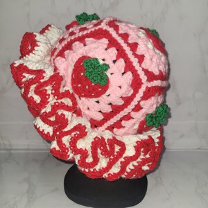 K&ouml;nnte beinhalten: Ein handgefertigter H&auml;kelhut im Erdbeer-Design. Der Hut hat ein rosa-rotes quadratisches Muster mit wei&szlig;en Akzenten und einem ger&uuml;schten Rand. Geh&auml;kelte Erdbeeren mit gr&uuml;nen Bl&auml;ttern sind am Hut befestigt.