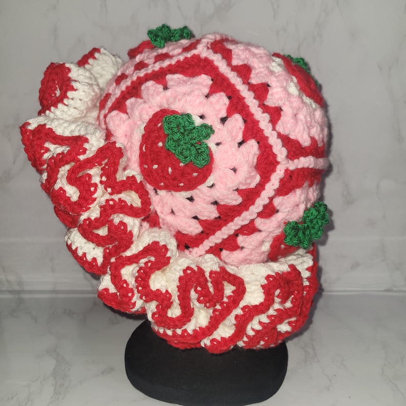 K&ouml;nnte beinhalten: Ein handgefertigter H&auml;kelhut im Erdbeer-Design. Der Hut hat ein rosa-rotes quadratisches Muster mit wei&szlig;en Akzenten und einem ger&uuml;schten Rand. Geh&auml;kelte Erdbeeren mit gr&uuml;nen Bl&auml;ttern sind am Hut befestigt.