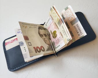 May include: A black leather wallet with a money clip holding several bills of Ukrainian hryvnia. The bills are folded and secured with the clip. The wallet is closed and has a smooth, textured surface.