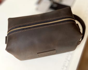 May include: A brown leather travel toiletry bag with a zipper closure and a small leather patch on the front.