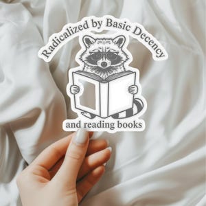 May include: A white sticker featuring a raccoon wearing glasses and reading a book. The text "Radicalized by Basic Decency" arches above the raccoon, with "and reading books" below.