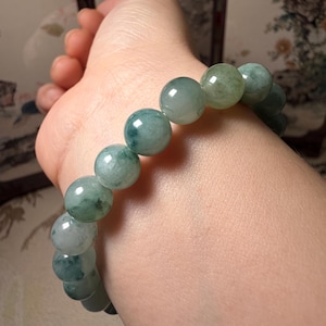 May include: A jade bracelet featuring large, round beads in shades of green and white. The beads are strung together to form a bracelet. The bracelet is shown on a wrist.