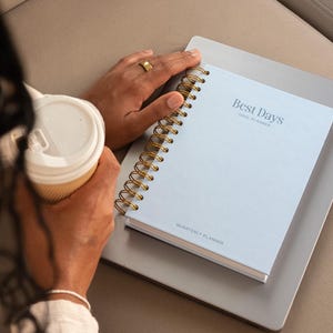May include: A light blue spiral-bound planner with gold-toned wire binding. The cover features the text "Best Days" and "Goal Planner" at the top, with "Quarterly Planner" printed at the bottom in a dark blue font.