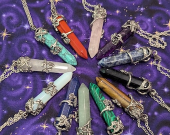 May include: A collection of gemstone pendant necklaces in various colors, including red, turquoise, pink, and green. Each pendant is a crystal point with a silver-colored metal floral design and chain. The necklaces are arranged on a purple background.