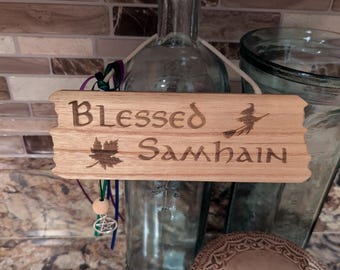 以下が含まれることがあります： 「Blessed Samhain」の文字が刻まれた木製の看板。ほうきに乗った魔女とカエデの葉も描かれています。コルク栓の付いた透明なガラス瓶の前に看板が吊り下げられています。
