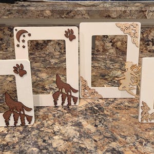 May include: Four white wooden picture frames with decorative cutouts. Two frames feature a wolf howling at the moon, paw prints, and stars. The other two frames have a fairy and floral corner accents. The frames are displayed on a speckled countertop.