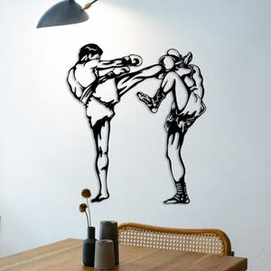 Könnte beinhalten: Schwarze Metall-Wandkunst, die zwei Figuren in einem Muay Thai-Kickboxkampf zeigt. Eine Figur tritt, die andere blockt. Das Kunstwerk ist an einer weißen Wand angebracht, mit einem Tisch und dekorativen Gegenständen im Vordergrund.