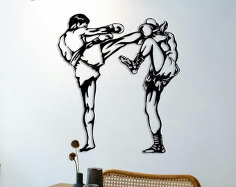 May include: Black metal wall art depicting two figures engaged in a Muay Thai kickboxing match. One figure is kicking, while the other is blocking. The artwork is mounted on a white wall, with a table and decorative items in the foreground.