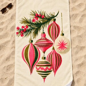 Top-down view of the whole retro Christmas ornament beach towel laid flat on sand, with plain cream space above and below the ornaments, a pine sprig, sunglasses and a small red gift box; one corner folds back to the white back.