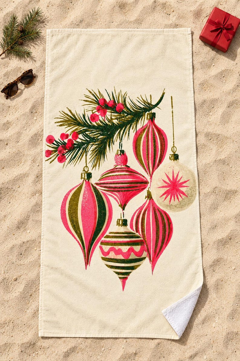 Top-down view of the whole retro Christmas ornament beach towel laid flat on sand, with plain cream space above and below the ornaments, a pine sprig, sunglasses and a small red gift box; one corner folds back to the white back.