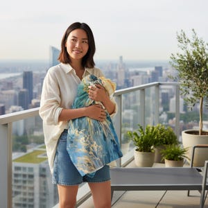 May include: A woman holding a blue, yellow, and white abstract patterned textile. The textile is wrapped in her arms. She is wearing a white button-down shirt and denim shorts. The background features a cityscape and a balcony.