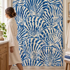 Blue scallop shell shower curtain in a bright bathroom with a wood vanity and woven rug, as a woman in a white robe walks past.