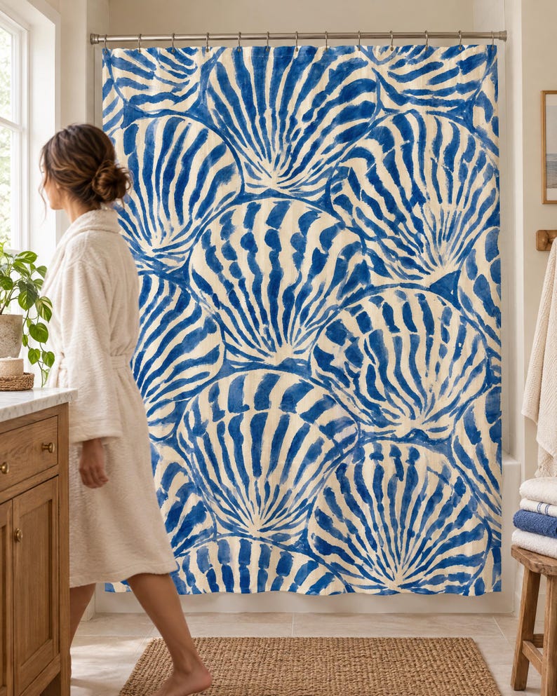 Blue scallop shell shower curtain in a bright bathroom with a wood vanity and woven rug, as a woman in a white robe walks past.