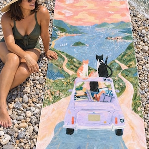 Cat road trip beach towel laid on a pebble beach, printed with a lilac car carrying cats and luggage down a winding road toward a pink-sky lake, as a woman in a green swimsuit and sun hat sits beside it.