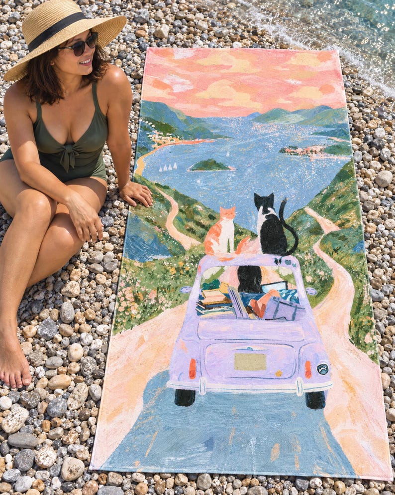 Cat road trip beach towel laid on a pebble beach, printed with a lilac car carrying cats and luggage down a winding road toward a pink-sky lake, as a woman in a green swimsuit and sun hat sits beside it.